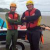 Photograph of two smiling members of Surf Life Saving New Zealand dressed in wetsuits and helmets. They stand infront of a jetski and its tralier which is parked on a beach