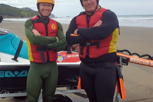 Photograph of two smiling members of Surf Life Saving New Zealand dressed in wetsuits and helmets. They stand infront of a jetski and its tralier which is parked on a beach