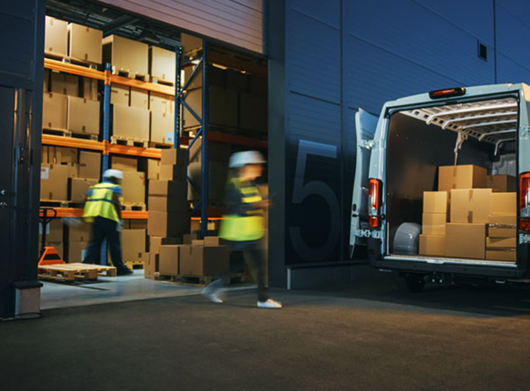 Photograph of two people wearing hard hats and safety vests loading boxes from a warehouse into a van.