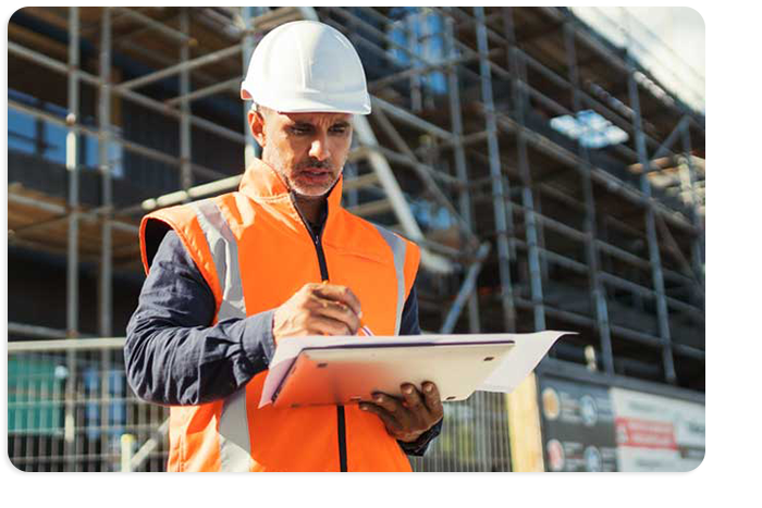Photograph of a contruction worker holding a laptop computer and paper document. Scaffolding and safety fencing of a building site can be seen in the background.