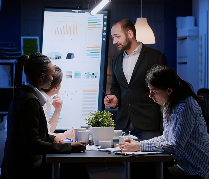 Three seated and one standing work colleagues around a table. A large vertical screen display data visulisations