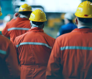 A photograph of multiple workers wearing orange safety suits and yellow hard hats seen from behind