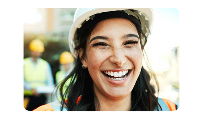A portrait photograph of a smiling worker wearing a hard hat and safety vest. The blurred background shows a further two colleagues also wearing hard hats and safety vests.
