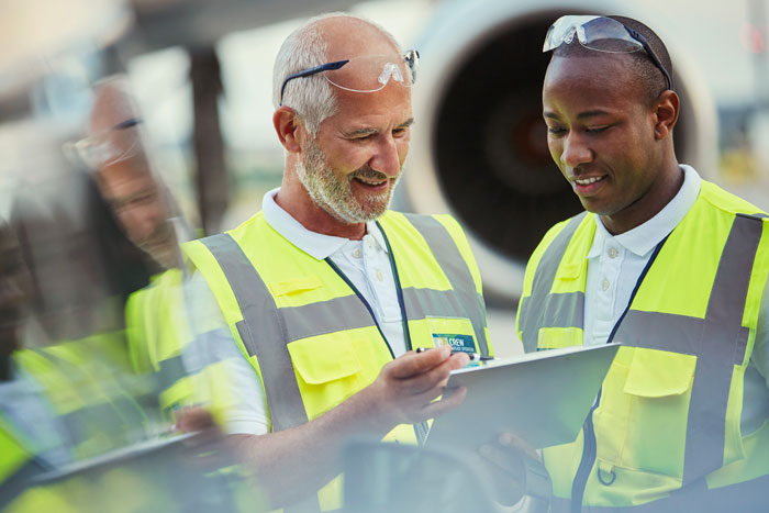 A photograph of two airport staff wearing high vibility vests examining a clipboard. In the background the engine of a large aircraft is visible