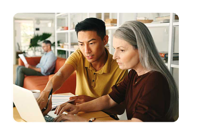 A photgraph of two people seated at a desk working from a laptop computer. Behind them a third colleague is seated on a couch reading a document