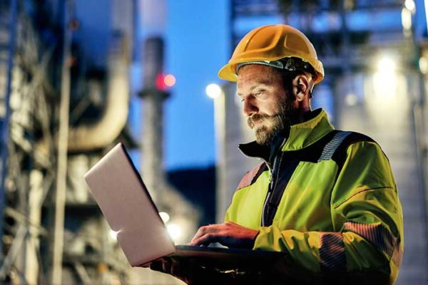 A person wearing a high visibility jacket and hard hat works on a laptop computer. The background shows a large industrial facility